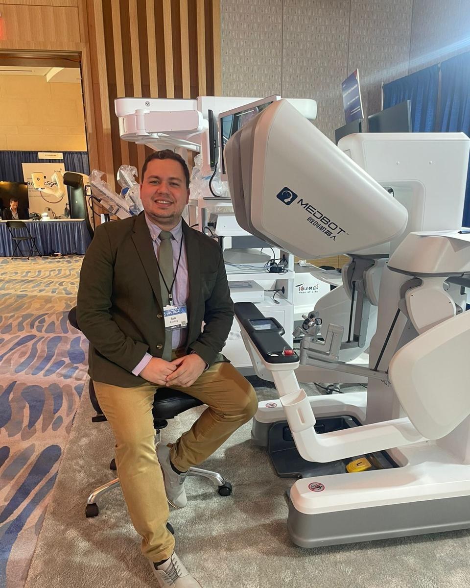 Dr. Ian Kauling ao lado de robô cirúrgico em congresso da Society of Robotic Surgeons